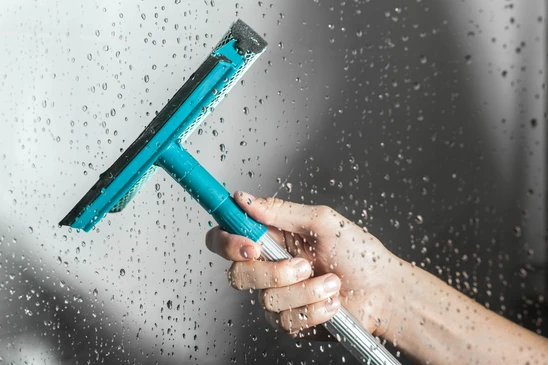 Une main tient une raclette de nettoyage turquoise sur une surface vitr&eacute;e couverte de gouttelettes d'eau