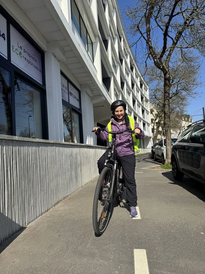 Gros plan d'un agent de nettoyage à vélo, équipé de sacoches noires professionnelles fixées sur le porte-bagages, portant un sac à dos ou une sacoche ventrale contenant du matériel de nettoyage adapté. L'agent est en tenue professionnelle (pantalon, polo ou chemise de travail), photographié dans une rue ou une résidence de Nantes avec des bâtiments résidentiels en arrière-plan. La composition doit montrer clairement le vélo comme mode de déplacement écologique et la préparation du matériel portatif, reflétant l'engagement écoresponsable de l'entreprise.